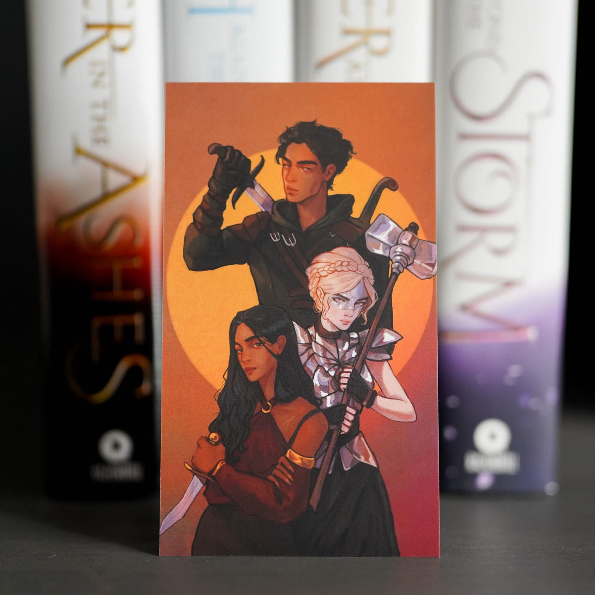 An Ember in the Ashes Adventure Card features Laia, Elias, and Helene holding weapons with character details on the back.