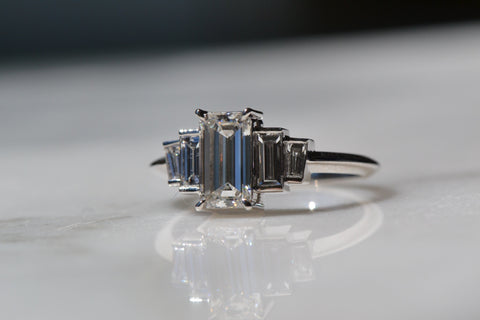 A close-up of an Art Deco Baguette Diamond Ring placed on the white shiny reflective surface.