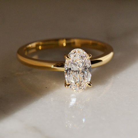 A close-up of an oval-cut  22k yellow gold ring with a diamond stone atop a shiny white surface.