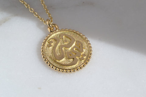 A close-up of a Gold " OM" Chain Pendant placed on a shiny surface.