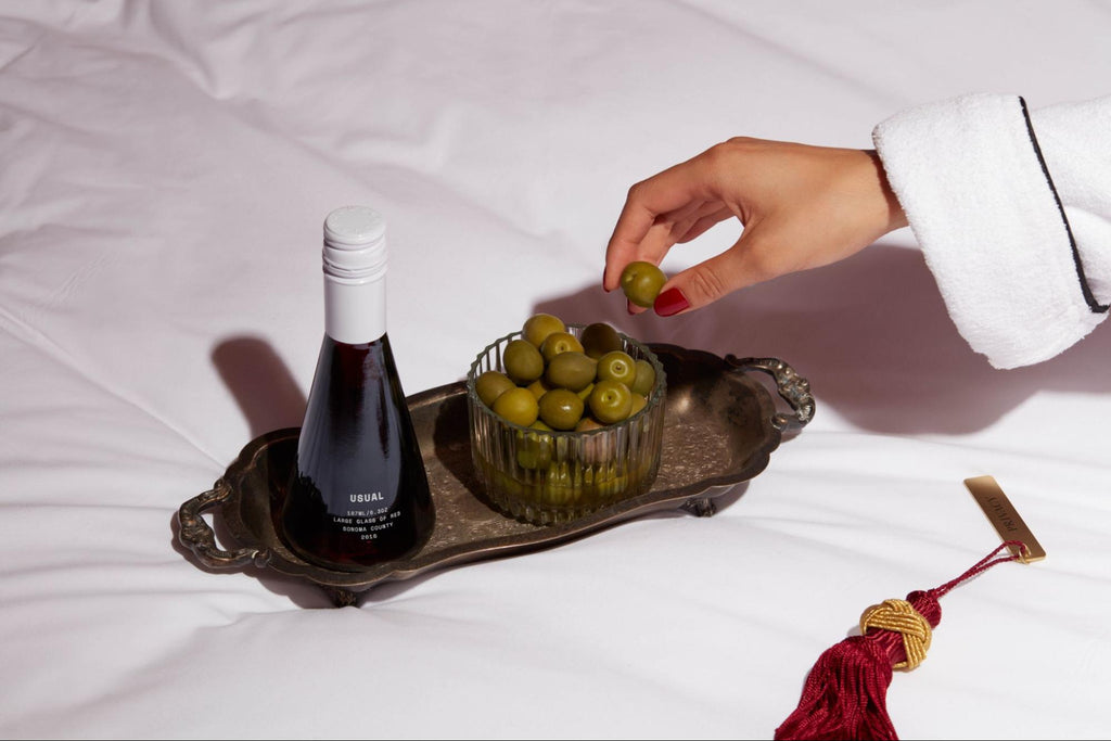 sweet red wine: person grabbing an olive from a bowl placed in a platter with a bottle of red wine from Usual Wines