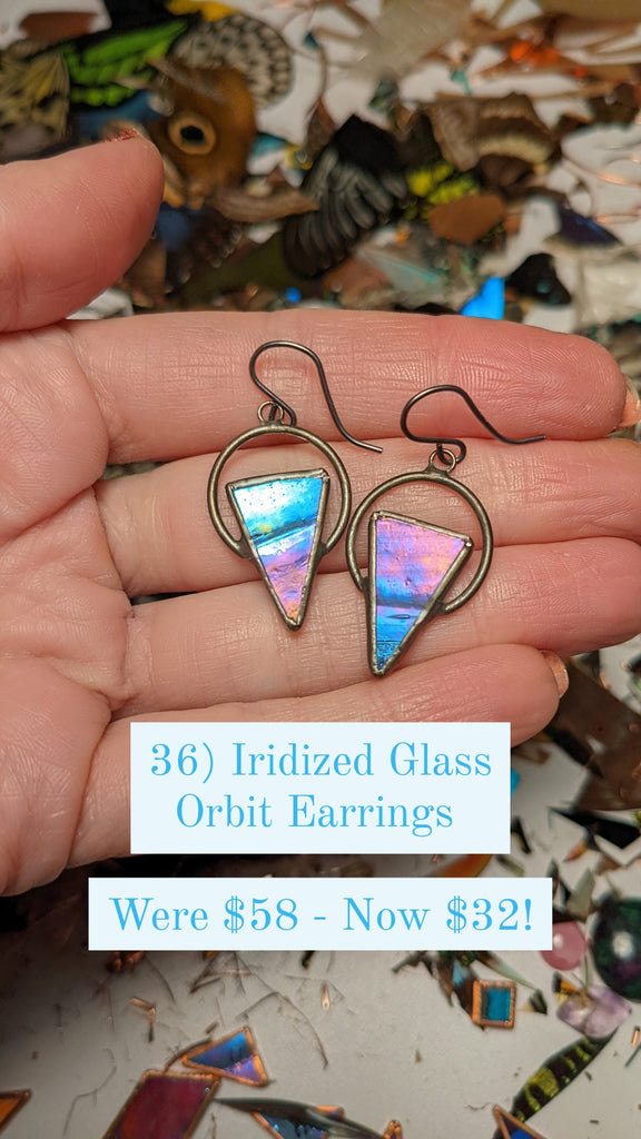 Mermaid colored iridized glass triangular earrings held in the palm of a woman's hand above a jeweler's workbench.