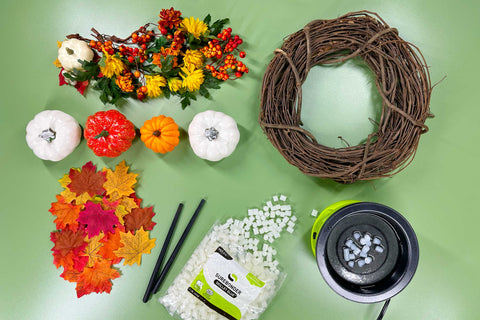 Supplies for project lay on a green tabletop. Fall leaves, small faux pumpkins in orange and white, floral sprays with orange berries and a grapevine wreath. Also included is the Surebonder glue skillet with melted glue inside, an open bag of low stringing glue pellets and two stir stick