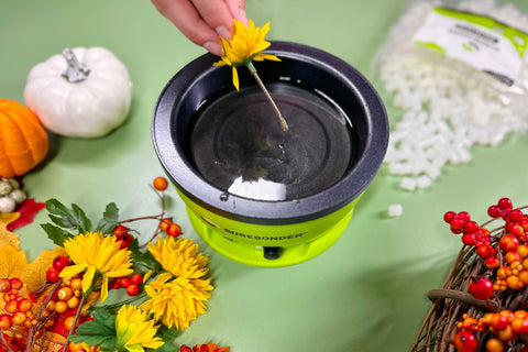 Surebonder glue skillet sits on a green tabletop surrounded by fall leaves, flowers, berry sprays and faux pumpkins. The stem of a yellow faux flower is being dipped into the glue in the skillet.