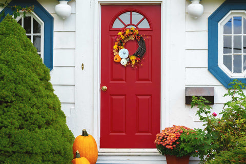 Front doorway entrance of a white house with a red door, pumpkins sit on the front steps. On the door is the completed project, a fall wreath in oranges and white in a crescent shape on a grapevine wreath