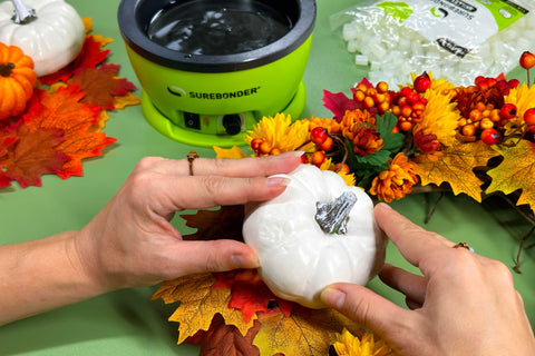 Hands are positioning and holding the faux white pumpkin onto the wreath that is filled with fall leaves, flowers and foliage.