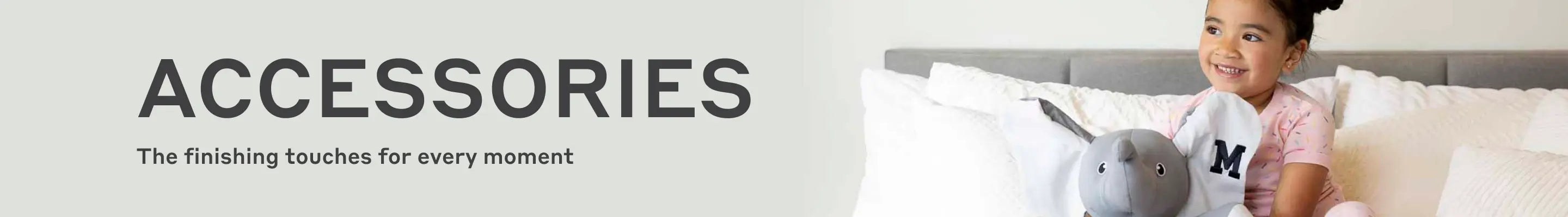Banner with text 'ACCESSORIES' and subtext 'The finishing touches for every moment', showing a smiling child sitting on a bed holding a gray elephant plush toy.