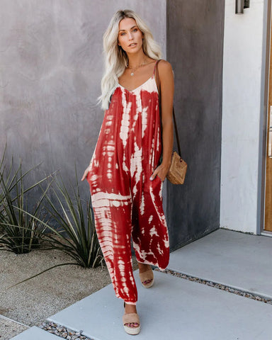 The image shows a stylish woman wearing a loose-fitting red-and-white tie-dye jumpsuit, paired with neutral espadrilles and a straw bag. She poses in front of a minimalist outdoor space with gray walls and decorative plants.