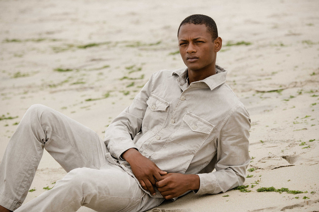 Lucas is reclining on the sandy beach, wearing twill shirt jacket and distressed button fly chino, both in khaki