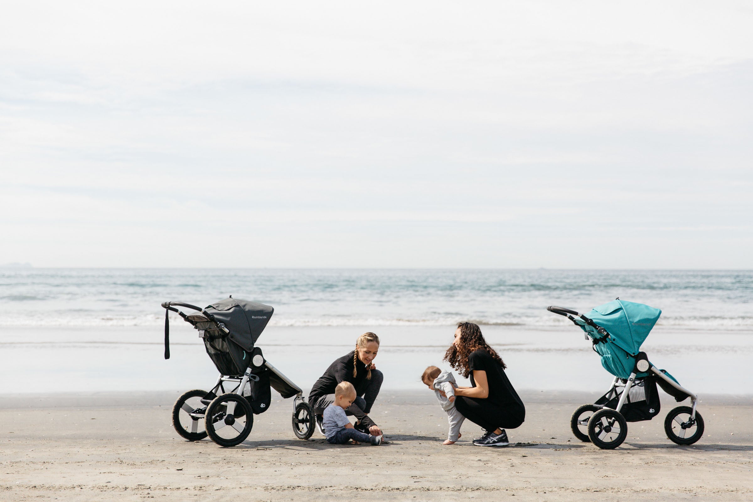 Picture of two Bumbleride families with their mothers and children kneeling on sand at beach at waters edge greeting each other