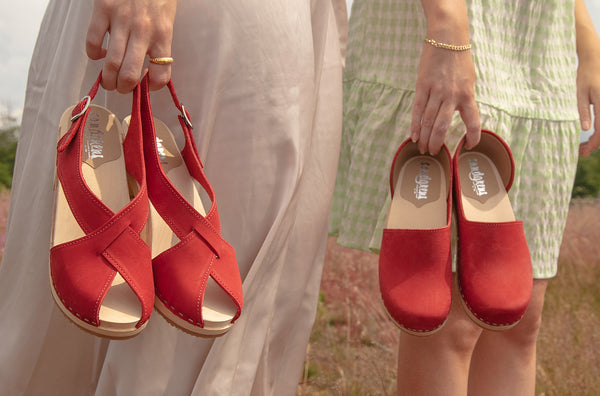 one girl holding up clog sandals in red leather, another girl holding up closed-back clogs in red leather