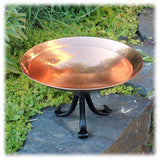 The shiny copper birdbath bowl is threaded onto a black wrought iron 3-legged base and displayed on a stone wall next to a bed of flowers