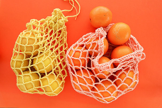 two bags of citrus fruit on a bright orange backdrop. The fruit are in crocheted reusable produce bags