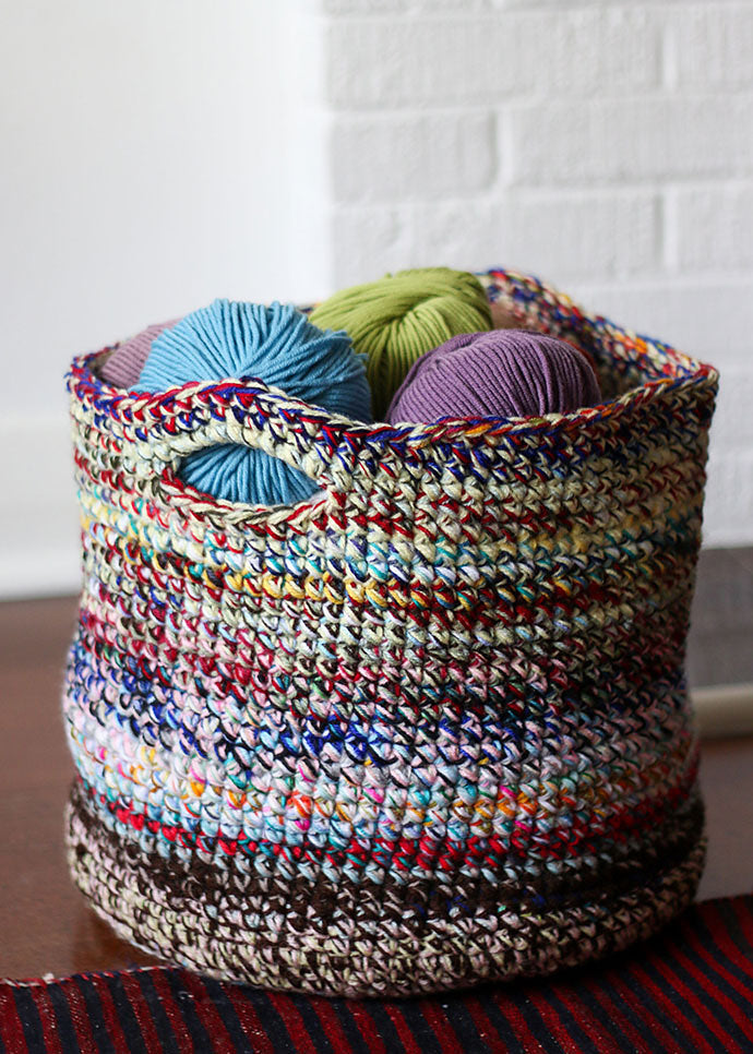 A colourful basket made of yarn is sitting on a wood table. Balls of wool fill the basket