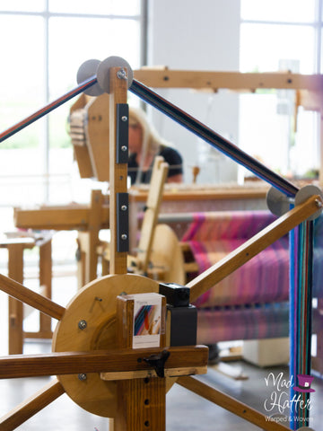 A wooden warping wheel with multicoloured yarn inside Mad Hatter Warped & Woven Studio. Wildflower can be seen weaving in the back in front of bright windows.