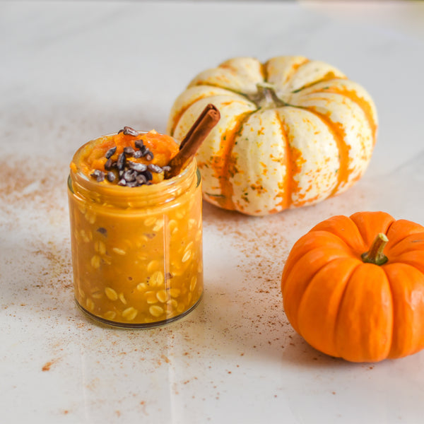 A small jar of pumpkin overnight oats sits on a white table next to two small pumpkins. 