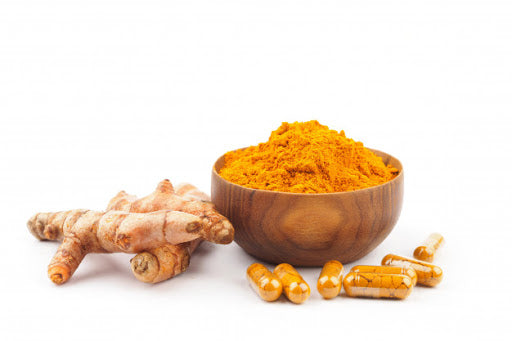 There is a bowl of turmeric powder next to tumeric root and supplements or curcumin on a white surface