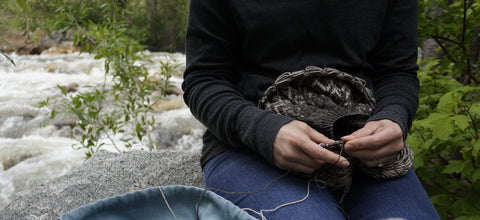 A closeup image woman shown knitting by a river. Only her midsection is visible, wearing dark clothing.