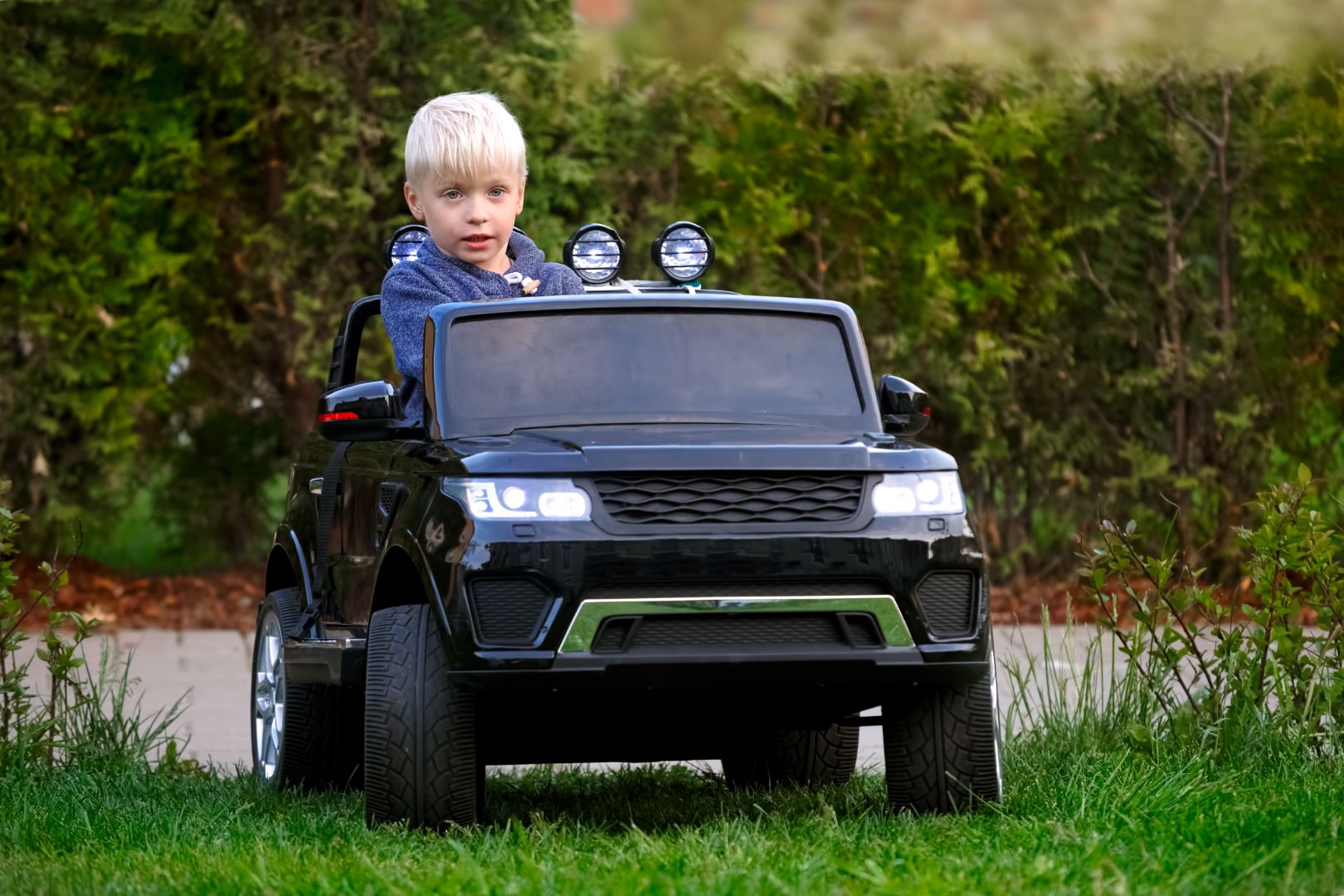 A blond child sits in his ride-on car in an area with tall grass, weeds, and a row of bushes behind him.