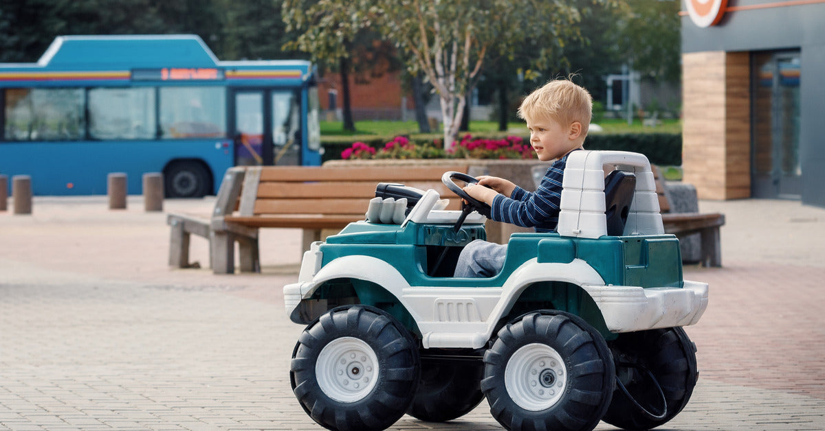 A young boy drives his green and tan power wheel through a public area. The child wears a blue-striped shirt.