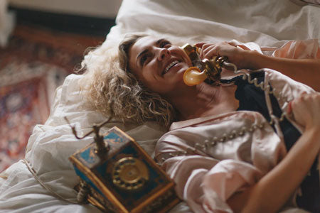 Smiling woman laying on a white bed while using a brown telephone.