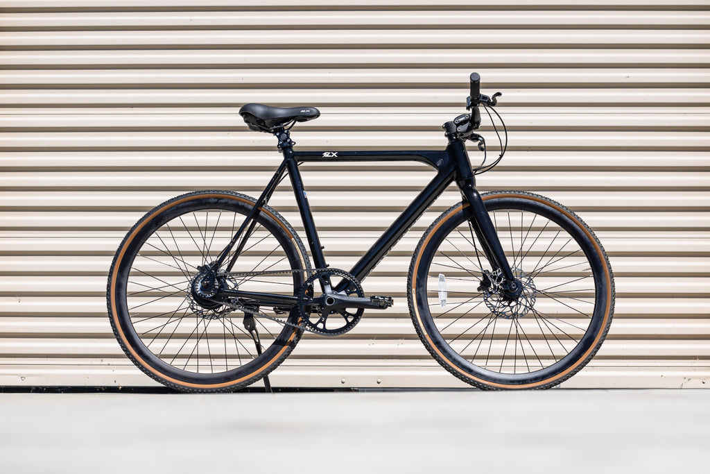 Black FLX Babymaker II steal electric commuter bike with tan wall gravel tires in front of a garage door in the sunlight