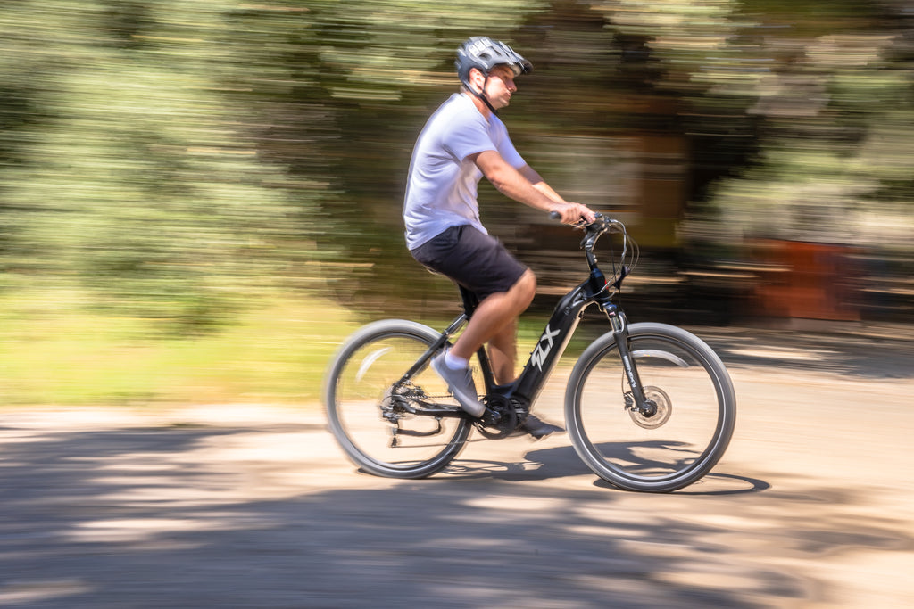panning photo of a young man riding an FLX Step Through electric comfort bike with upgraded gravel tires on an off road trail