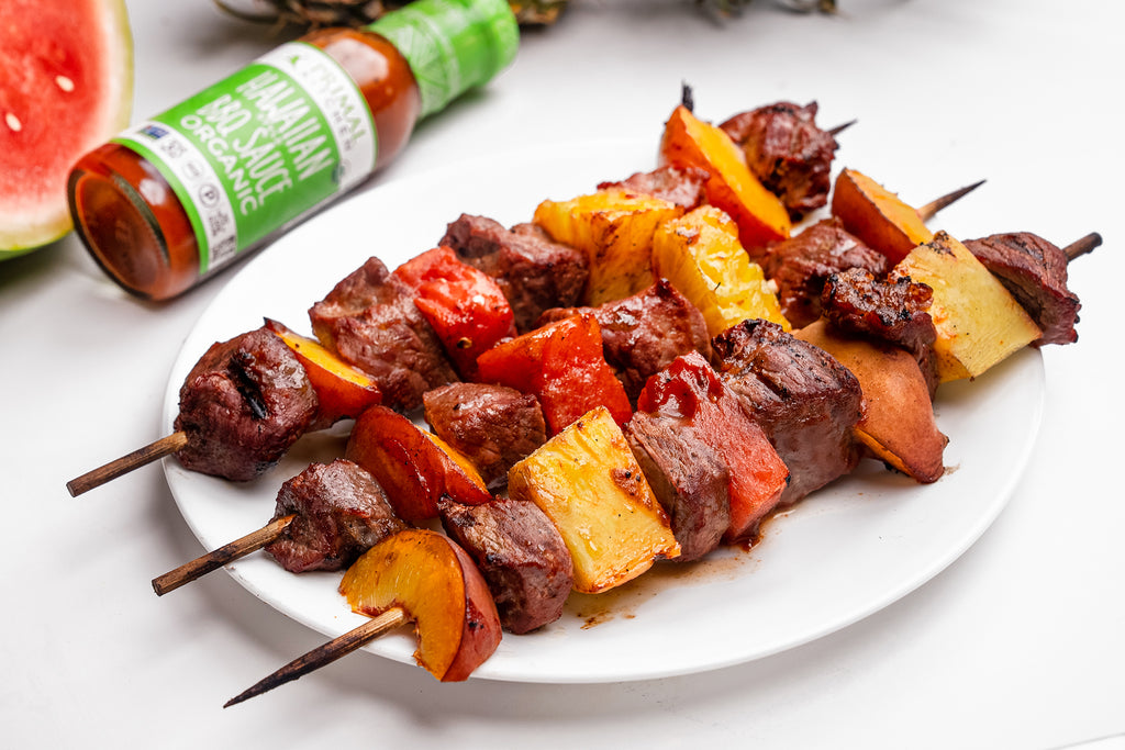 A tray of Hawaiian steak and fruit kabobs next to a bottle of Primal Kitchen Hawaiian Style BBQ Sauce and Marinade