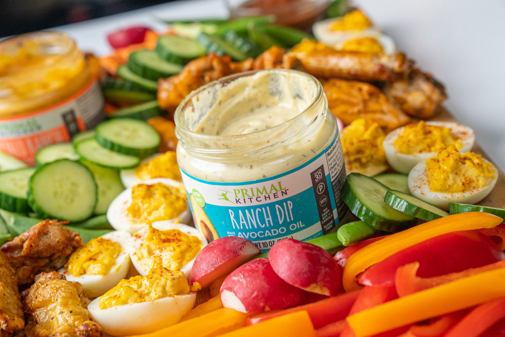 Closeup of Primal Kitchen Ranch Dip surrounded by fresh veggies, wings, and deviled eggs on a wooden board.