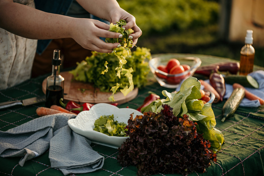 Hands tearing lettuce for a salad on an outdoor table with assorted fresh vegetables.