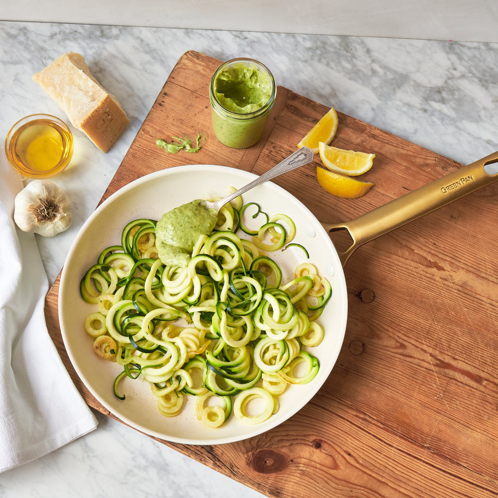 zucchini noodles in a pan on a wood cutting board with homemade pesto sauce in the back