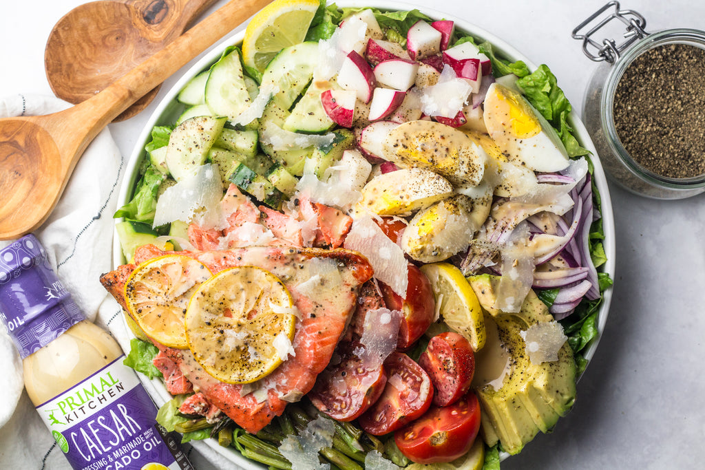 A large bowl of baked salmon caesar salad next to a bottle of Primal Kitchen Caesar Salad Dressing