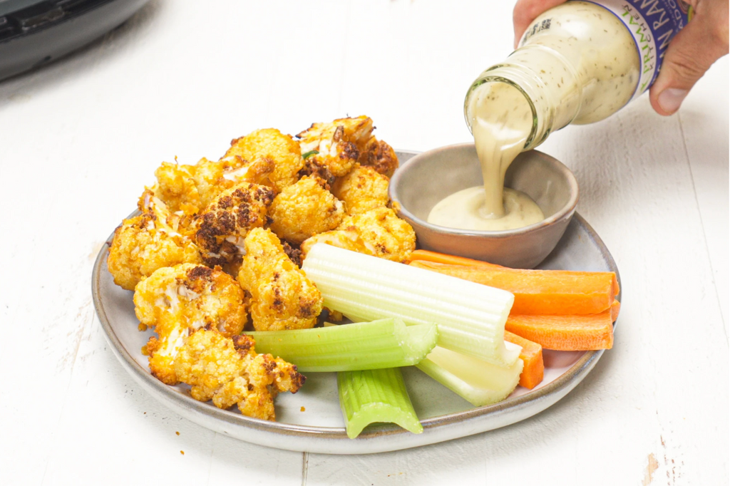 A hand pours ranch into a dish with buffalo cauilflower and carrots and celery sticks nearby.