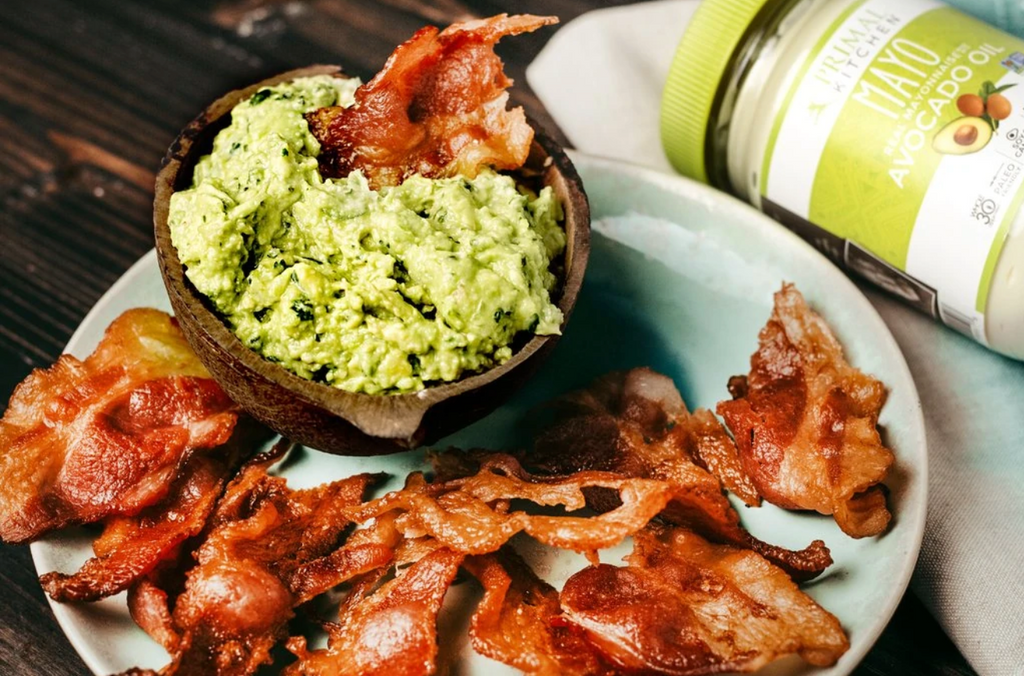 A strip of bacon is dipped in gaucamole on a plate of bacon with primal kitchen mayo behind it.