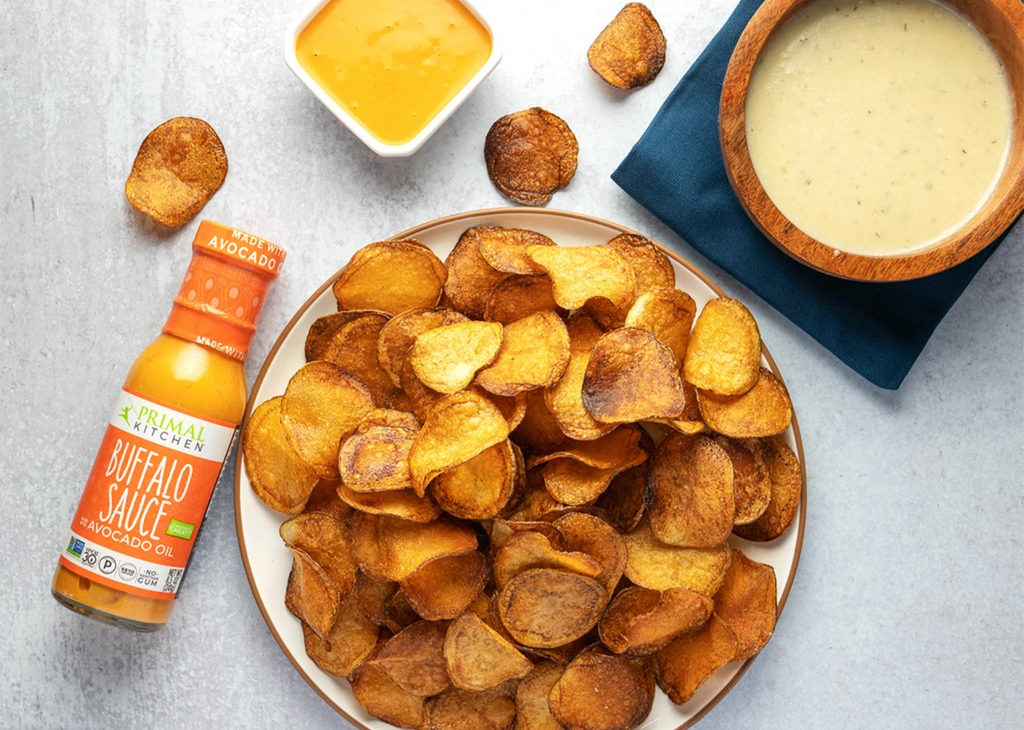 Homemade potato chips are arranged on a plate with two dipping sauces and a bottle of buffalo nearby.