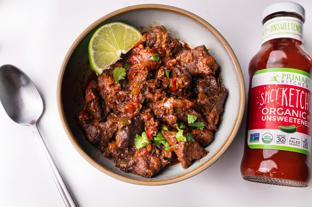 Spicy ketchup and a silver spoon are laying next to a bowl of chili.