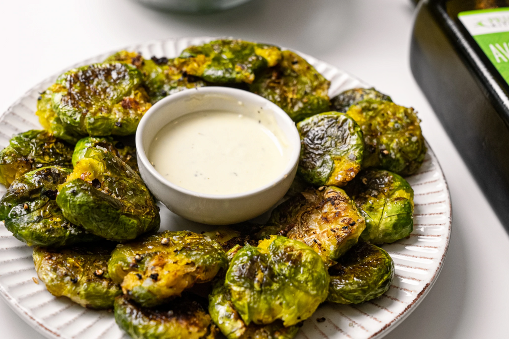 Smashed brussel sprouts are arranged around ranch dip on a white plate with avocado oil off to the side.