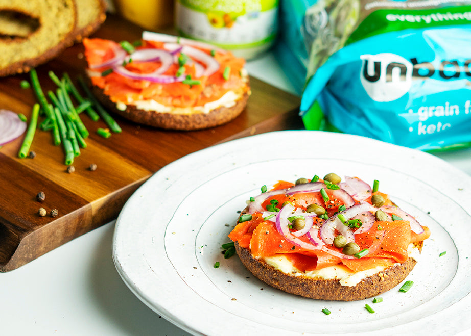 Smoked salmon bagel on a white plate with another bagel half on a white board, and bread package in the background.