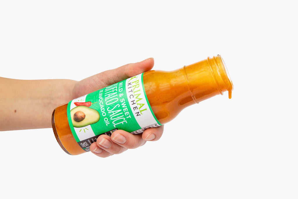 A hand holding a bottle of Primal Kitchen Mild Buffalo Sauce, with a drizzle of sauce coming out.