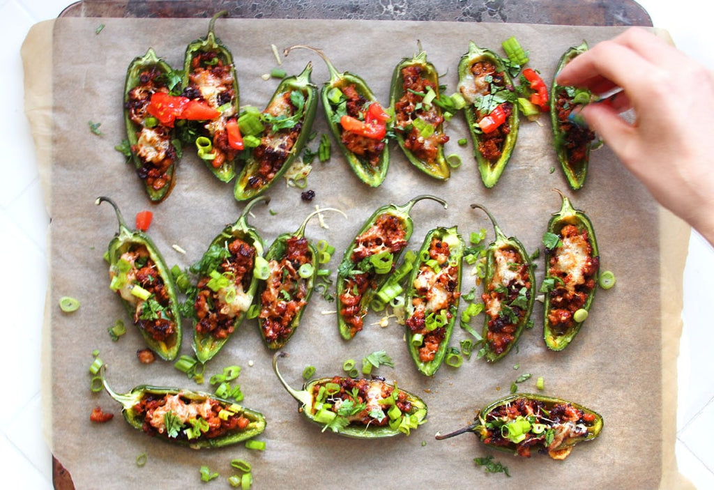 A hand sprinkling garnishes over a tray of jalapeno stuffed peppers