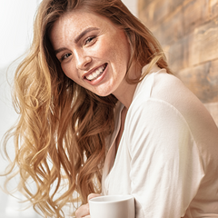 Beautiful woman with long wavy hair, freckles and full eyebrows wearing a robe and holding a coffee cup.