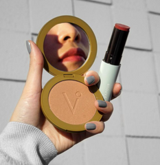 woman's lips reflected in bronzer powder mirror she's holding, also holding Vapour Multi Stick. The woman is wearing grey nail polish. 