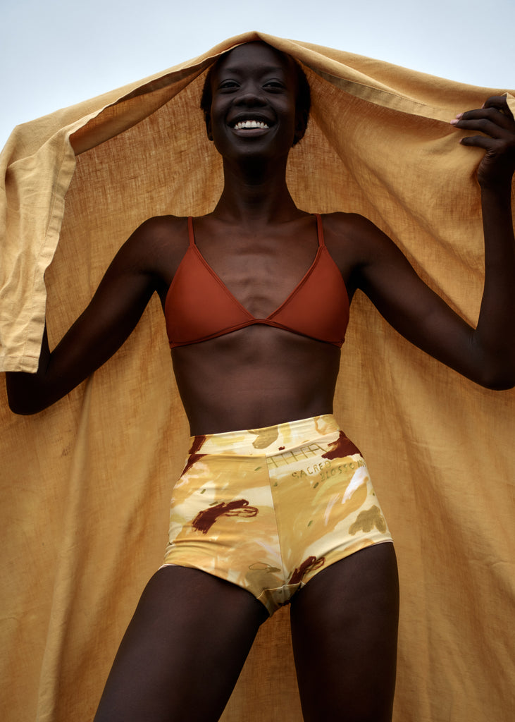 hakea x studio of the sun yellow abstract floral print bikini short with rust red triangle bikini top and mustard yellow towel held up behind model