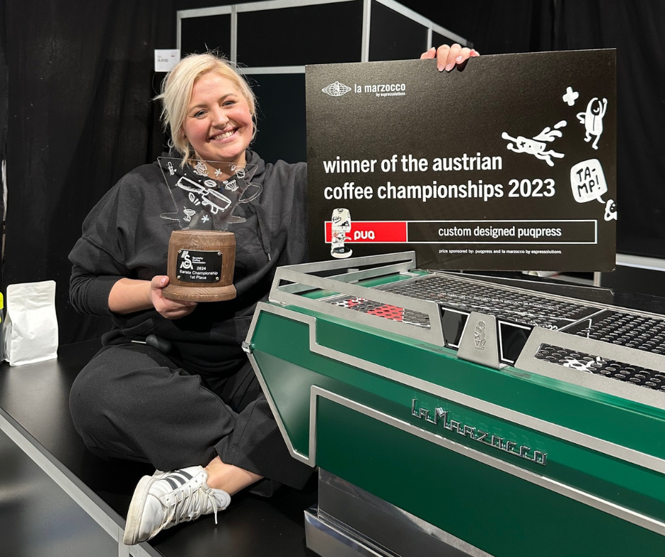 Barista Dominika Piotrowska sitting beside a green espresso machine and a sign proclaiming her the winner of the Austrian Barista Championships 2023