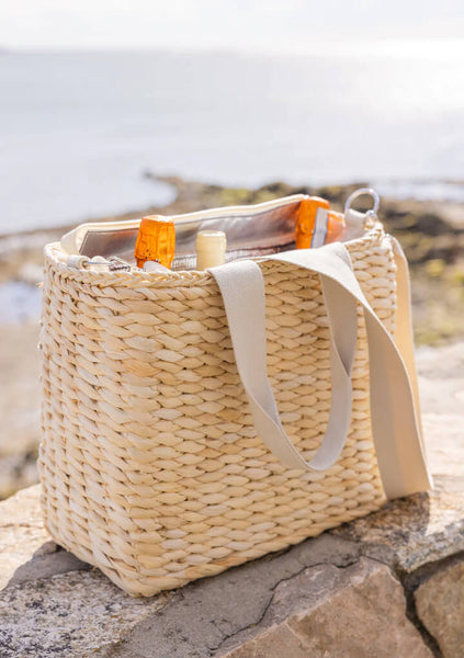 Straw beach bag with bottles and towels on rocks.