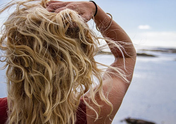 Back view of a woman's curly blonde hair, hand raised, at the seaside.