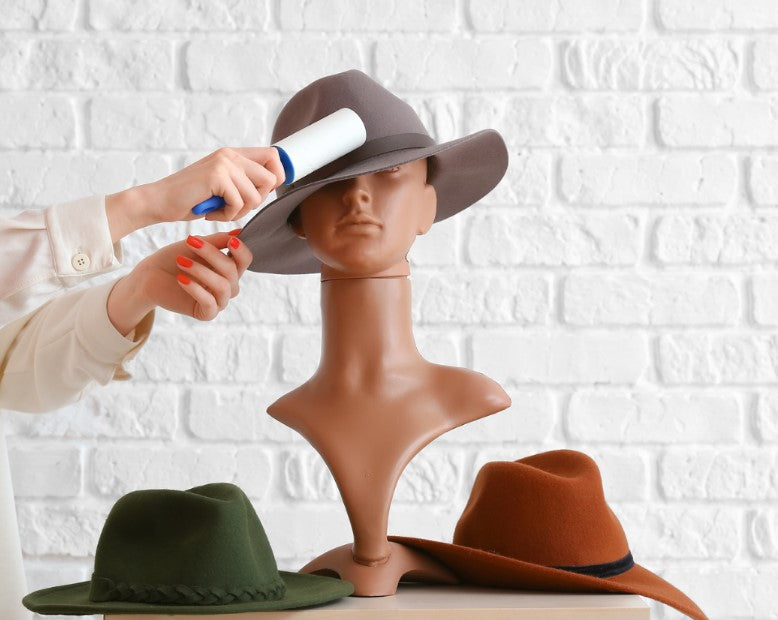 A hand using a lint roller on a brown fedora hat placed on a mannequin head, with two other hats (one green and one rust-colored) and a white brick wall in the background.