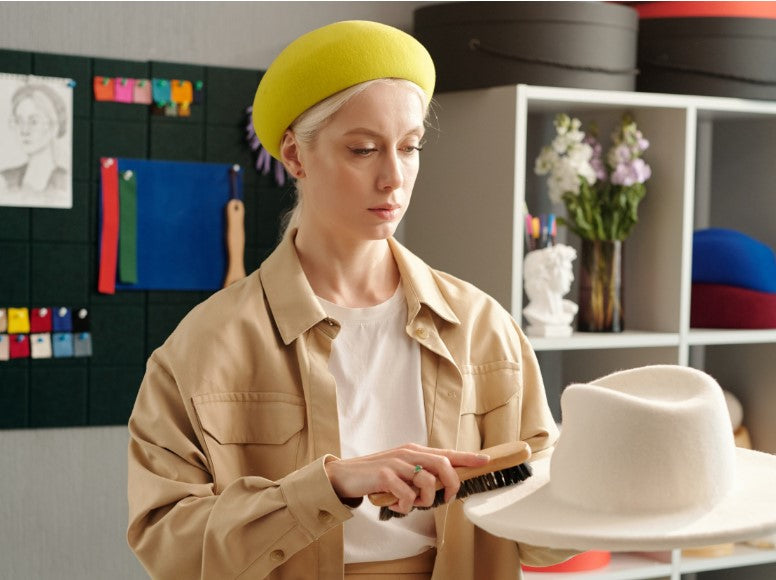 Blond woman looking down while brushing a white fedora hat with a small brush.