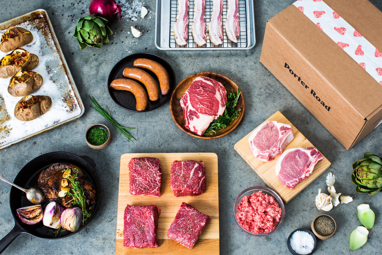 Porter Road meat subscription box shown with a display of steak, sausage, bacon, baked potatoes, and other ingredients.