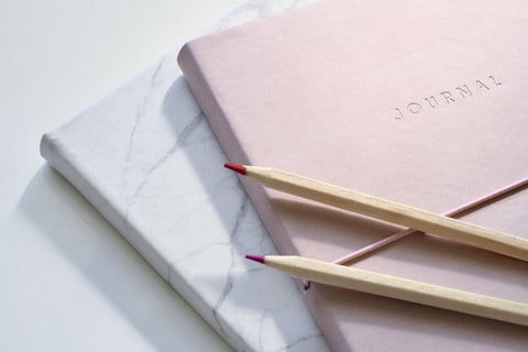 two beautiful mindfulness and gratitude journals on top of each other on a white surface or desk, one has a pink cover the other a blue cover and there are two pencils on top of them